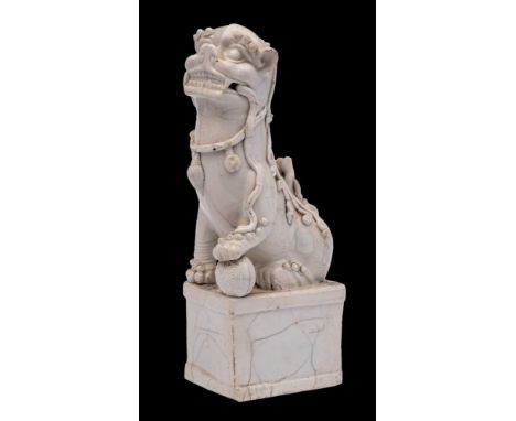 A Chinese blanc-de-chine Buddhist lion dog: on raised rectangular plinth, one paw resting on a brocade ball, a ribbon held in