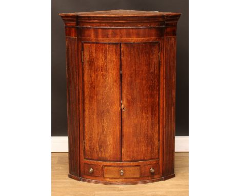 A George III mahogany crossbanded oak bowfront corner cupboard, moulded cornice above a pair of doors enclosing two shelves, 