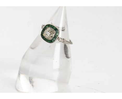 An emerald and diamond Art Deco style dress ring, the cushion cut diamond surrounded by a band of calibre set square cut emer