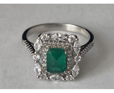 A WHITE METAL RING WITH A CENTRE RECTANGULAR LABORATORY GROWN EMERALD SURROUNDED BY CLEAR STONES SIZE Q/R 