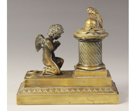 A brass desk inkwell, 19th century, designed as a cherub kneeling before a memorial plinth, surmounted by a sword and helmet,