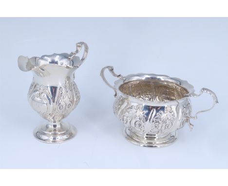 A George V silver cream jug and sugar basin, respectively of helmet and ovoid form, both repoussé decorated with repeating fl