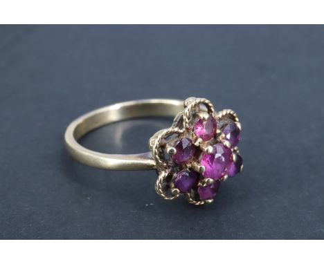 A ruby daisy ring, having a central 4 mm brilliant set above six surrounding 3 mm brilliants, their outline traced by a fine 
