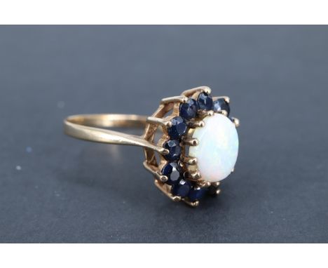 An opal and blue sapphire finger ring, the 8 x 6 mm opal cabochon set above 12 surrounding sapphire brilliants, each of 2.5 m