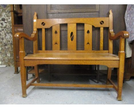 An Arts & Crafts oak bench having a splat back with heart and arrow pierced ornament, curved arms and solid seat, raised on s