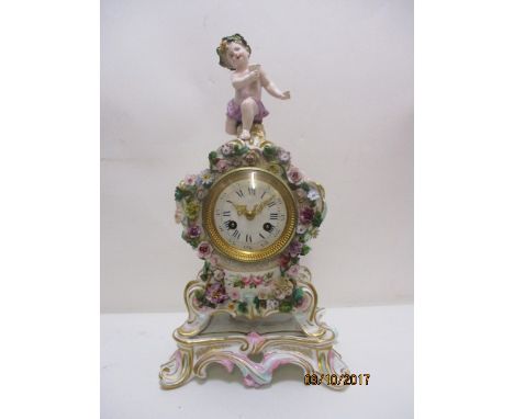 A late 19th century German porcelain cased mantle clock, encrusted with flowers and foliage, surmounted by a cherub on an att