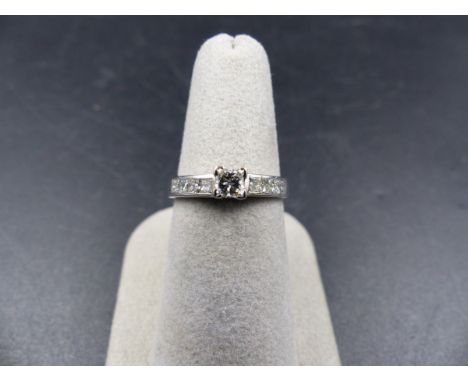 A WHITE METAL DIAMOND RING. THE CENTRAL DIAMOND IS A PRINCESS CUT IN A FOUR CLAW CORNER SETTING WITH PRINCESS CUT CHANNEL SET