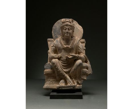 100 – 300 AD. Gandharan. Schist figure of a seated Buddha atop a throne backed by a halo. Buddha wears an elaborate headpiece