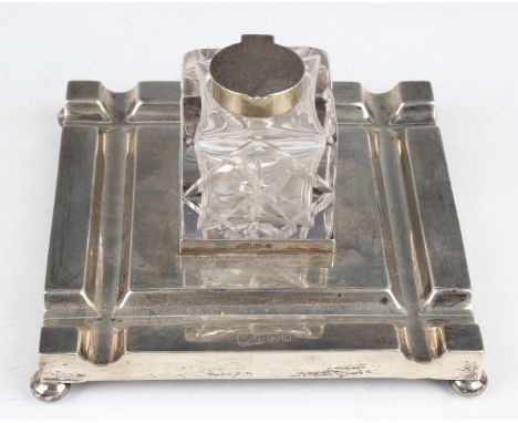 A George V silver square inkstand, fitted with a central cut glass inkwell with silver collar and hinged lid, on bun feet, Bi