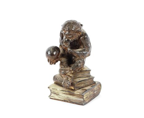 A silvered novelty table lighter, in the form of a seated monkey with hinged head, raised on a pile of books and holding a sk