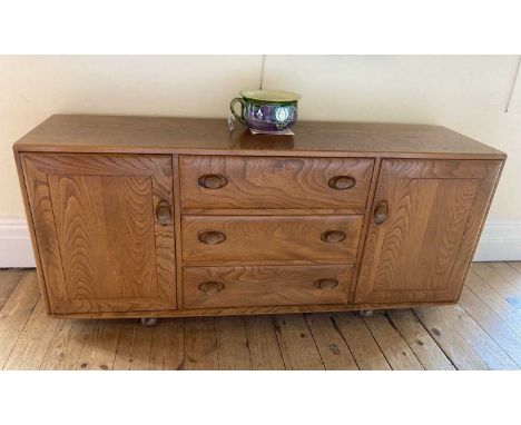 AN ERCOL LIGHT ELM SIDEBOARD, with three central drawers flanked on either side by a cupboard door, oval wooden handles on ca