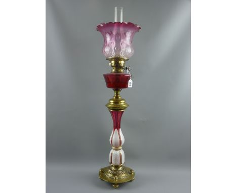 A Victorian oil lamp with circular embossed brass base on four brass feet, white overlay and ruby cut glass stem, central rub