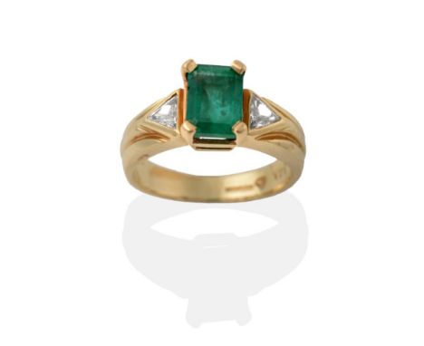 A 14 Carat Gold Emerald and Diamond Ring, the step-cut emerald in a yellow claw setting, flanked by trilliant cut diamond cha