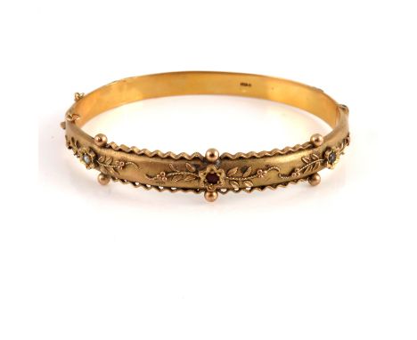 An Edwardian 9ct yellow gold hinged bangle, raised floral decoration inset with gemstones and pearl, 8.1gms 