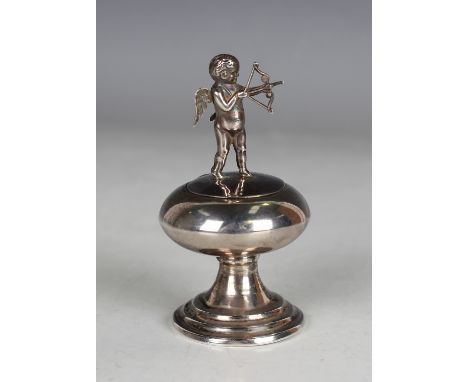 A George V silver trinket box, the hinged lid surmounted by Cupid firing an arrow above a flattened circular body, raised on 