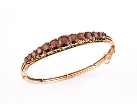 A GARNET AND GOLD HALF HINGED BANGLE mounted to one side with graduated oval-shaped garnets, internal measurements 6.2 x 5.5c