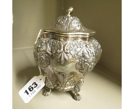A silver repousse tea caddy, finial to top over a hinged lid, all over floral decoration standing on four paw feet, hallmarke