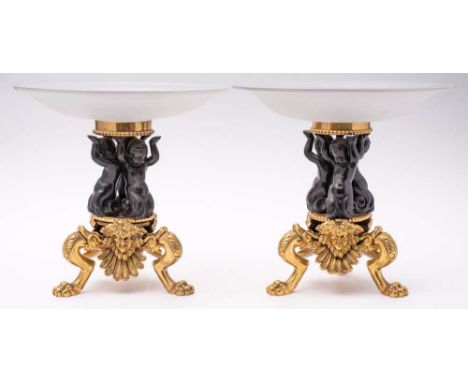 A pair of bronze and gilt bronze tazze: the opaque circular glass dishes mounted on three cherub supports to a circular plint