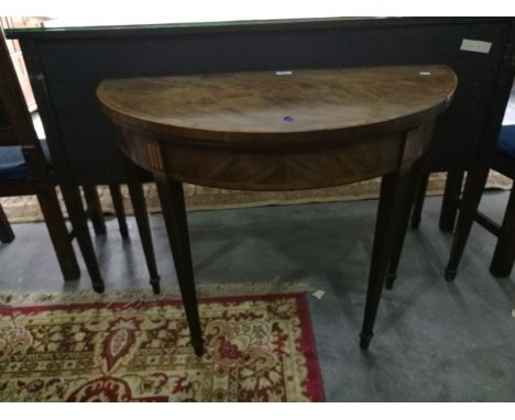 A figured mahogany demi-lune card table on reeded tapering supports (the back leg is coming away, old marks and scratches to 