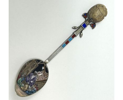 A silver coloured metal and enamel teaspoon, decorated in the Egyptian manner, the finial inlaid with a carved stone scarab b