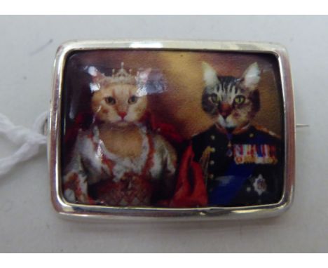A silver coloured metal framed brooch, set with an enamel tablet, depicting two Royal Cats 