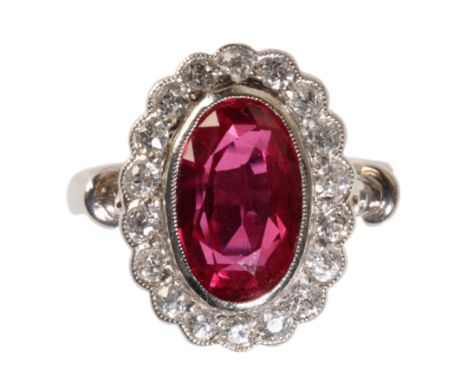 A SYNTHETIC RUBY AND DIAMOND CLUSTER RING, the oval-cut ruby, 11mm x 7mm, collet-set within a border of eighteen brilliant-cu