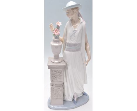 A Lladro porcelain figurine of a 1920's flapper girl modelled next to a jardiniere stand. Stamped Lladro to the base. Measure