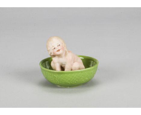 A small Gebruder Heubach bathing girl,  seated leaning forward with hand between feet, green glazed basin and impressed starb