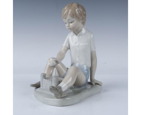 Glossy porcelain figurine that features a young boy sitting as he carefully builds a sandcastle. This piece details a serene,
