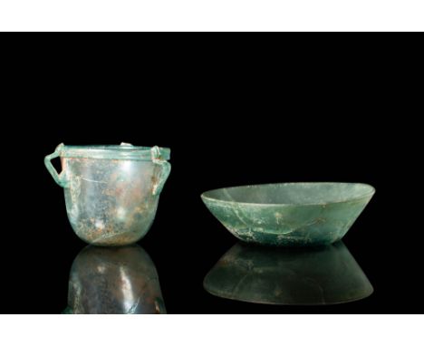 Ca. AD 100 - 300 .
A pair of glass vessels. The first has a cup-shaped body with a folded rim and strap handles. The second i