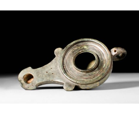 Ca. AD 100 - 300.
A cast-bronze oil lamp. The globular-shaped body has a rounded spout adorned with stylised volutes, raised 