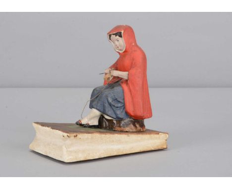 A mid 19th century Thüringen papier-mâché girl knitting squeak toy,  the girl with red cloak, seated on a rock with moving ha
