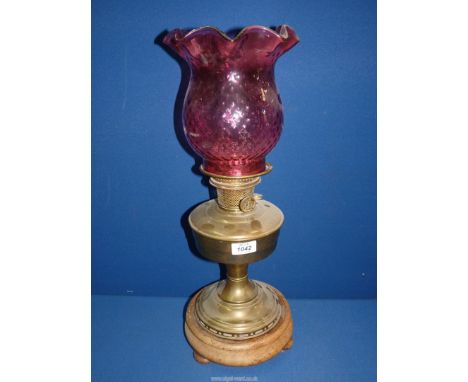 A Brass oil lamp on a wooden base having three bun feet with a pretty cranberry shade.