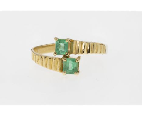 An 18ct gold and emerald ring, featuring two step cut emeralds held in a crossover setting on a reeded shank, ring size P1/2 