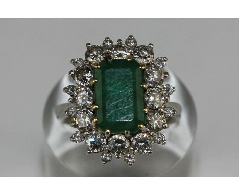 An emerald and diamond cluster ring, the step cut stone set within a two row border of brilliant cut diamonds in 18ct white g