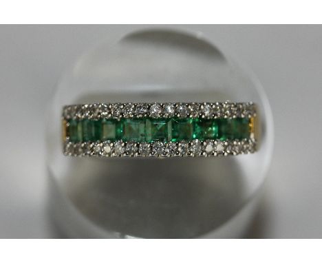 An emerald and diamond ring channel set with a band of nine square cut emeralds between diamond borders on 18ct gold