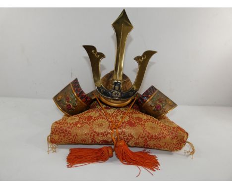 A 20TH CENTURY JAPANESE MINIATURE KABUTO HELMET WITH CAST BRASS DECORATION, TASSELS, MOUNTED ON A DECORATIVE CUSHION. HEIGHT 