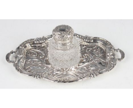 A late Victorian silver inkstand of shaped oval two-handled form, embossed with scrolling leaves against a textured ground wi