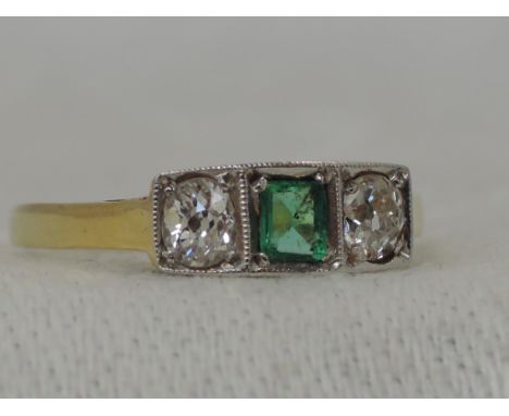 A lady's dress ring having a central step cut emerald flanked by two old cut diamonds in a rectangular pave set mount on a ye