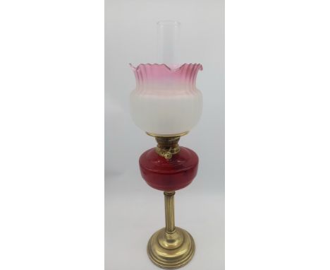 An antique brass column base oil lamp with cranberry glass reservoir with shade and chimney. 