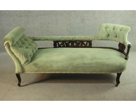 An Edwardian chaise longue, upholstered in green buttoned velour, the back panel carved and pierced with a stylized foliate d