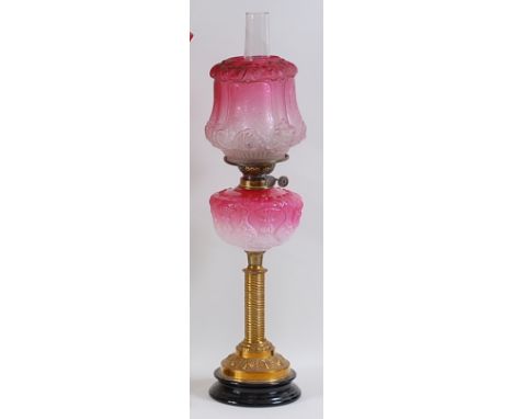A late Victorian brass pedestal oil lamp, having a cranberry tinted and acid etched shade, above a moulded shaded pink glass 