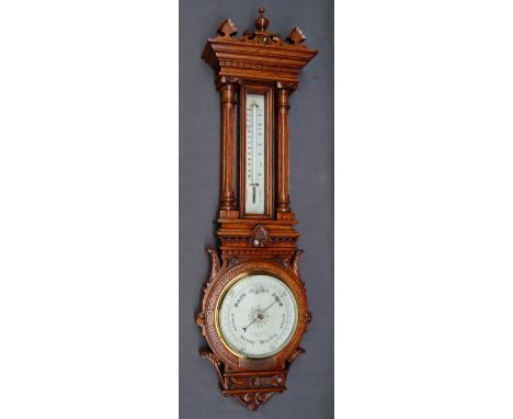 An Edwardian oak aneroid barometer with a carved and turned cresting rail above a mercury thermometer, the silvered dial insc
