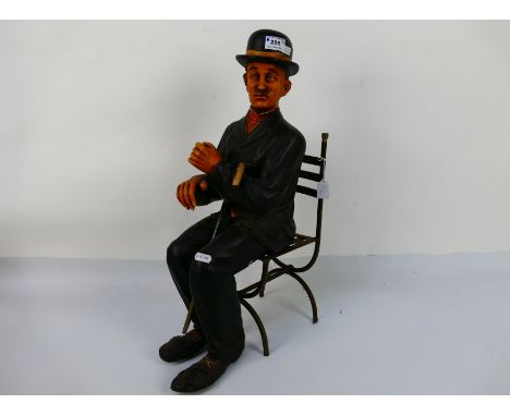 A figure depicting Charlie Chaplin in a seated pose with metal chair, approximately 57 cm (h). [2]. [W]. - Condition report -