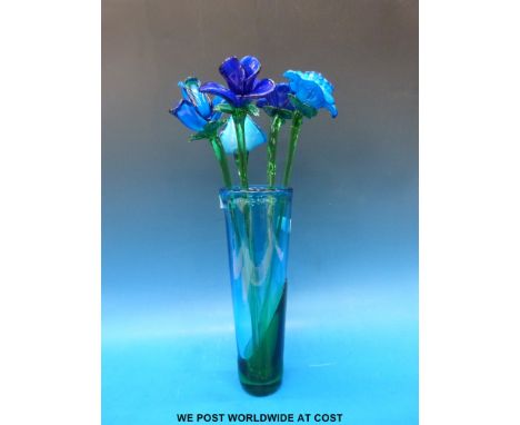 A blue-green glass vase containing five blue glass flowers with green stems