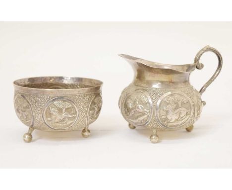Indian white-metal milk jug and sugar basin, each decorated with panels of animals in landscapes and standing on three ball f