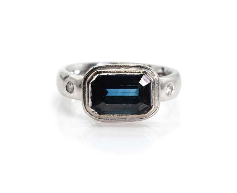A platinum sapphire and diamond ring,  with a central emerald cut sapphire, bezel set with a wire surround, into shoulders ea