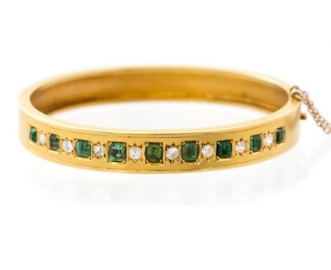 An Edwardian emerald and diamond set 18ct yellow gold hinged bangle, a total of eight emerald cut emeralds alternating with e