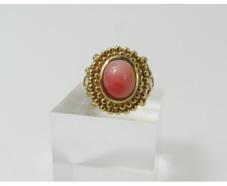 A 9ct gold pink stone dress ring, the oval cabochon cut stone (probably coral), in a rub over setting, with two rows of beadi