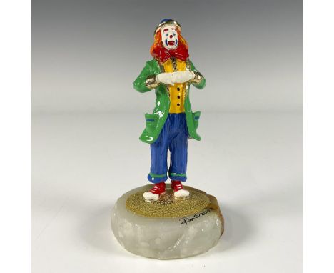 A gilded enamel figure of a smiling clown wearing a green jacket, large red bow tie, and blue pants. Presented on a white ony